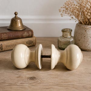 May include: A pair of cream-coloured antique door knobs with a classic round design. The knobs are separated, showing the connecting hardware. A small brass bell and vintage books are in the background, suggesting a vintage aesthetic.