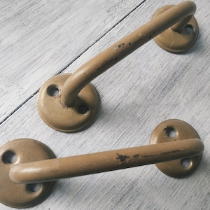 May include: Two vintage-style, tan-colored metal handles. Each handle has a rounded grip and a circular base with two screw holes. The handles show signs of wear, with some paint chipping, suggesting an antique or rustic aesthetic.