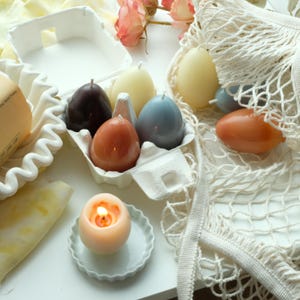 May include: Egg-shaped candles in various colors, including brown, orange, and blue, are displayed in an egg carton and a net bag. A lit candle sits on a white plate. Pink roses and a yellow fabric add to the aesthetic.