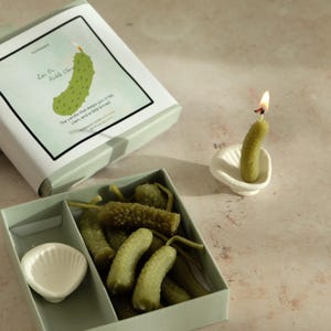 May include: A set of pickle-shaped candles, some in a light green box, and one lit in a white seashell-shaped holder. The box reads "Zen On Pickle Clam". The lit candle has a small flame.