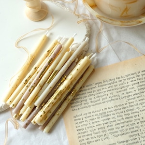 Könnte beinhalten: Ein Bündel dünner, weißer Kerzen mit goldenen und braunen Akzenten. Die Kerzen sind mit goldenen Flocken verziert und haben eine rustikale, vintage Ästhetik.