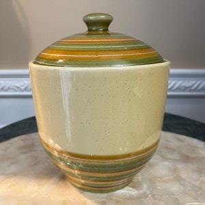 May include: A ceramic canister with a lid, featuring a striped pattern in shades of green, brown, and white. The canister has a speckled, cream-colored body.
