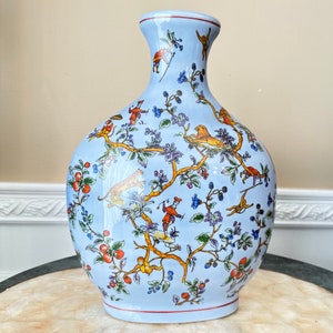 May include: A light blue ceramic vase with a floral and animal design. The vase has a wide base and a narrow neck. The design features a variety of flowers, birds, and animals in shades of orange, yellow, and green.