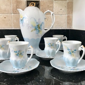 Può includere: Un set da tè in porcellana vintage con un designo floreale. Il set comprende una teiera, sei tazze e sei piattini. Le tazze e i piattini sono decorati con delicati fiori blu e un bordo blu chiaro. La teiera ha un manico dorato e un designo floreale sul corpo.