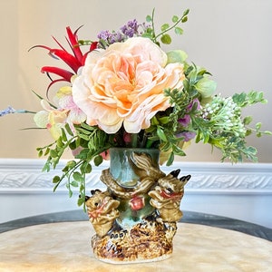 Puede incluir: Un jarrón de cerámica con un esmalte verde y un diseño de dragón marrón y beige. El jarrón está lleno de un ramo de flores melocotón, rosa y blancas.