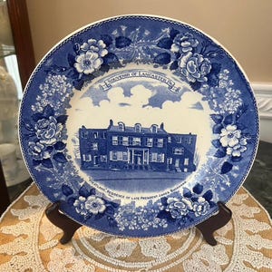 May include: Blue and white decorative plate featuring a detailed illustration of a building, surrounded by floral patterns. The plate has the text "Souvenir of Lancaster, PA" and "Wheatland, Residence of Late President James Buchanan".