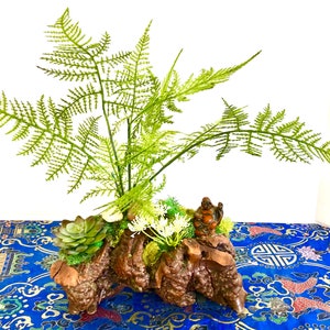 以下が含まれることがあります： 青い花柄の布の上に置かれた、木の鉢植えの緑色のシダ植物。鉢には小さな多肉植物と、岩に座っている人の小さな置物が置かれている。