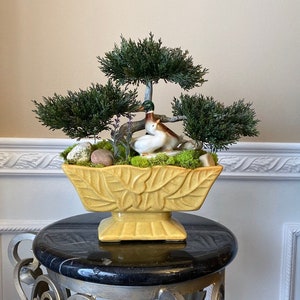 May include: A yellow ceramic planter with a leaf design, filled with moss, small rocks, and a miniature bonsai tree with two ceramic ducks nestled in the greenery.
