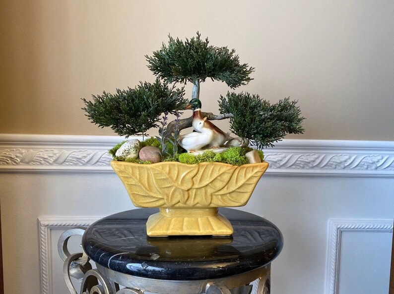 May include: A yellow ceramic planter with a leaf design, filled with moss, small rocks, and a miniature bonsai tree with two ceramic ducks nestled in the greenery.