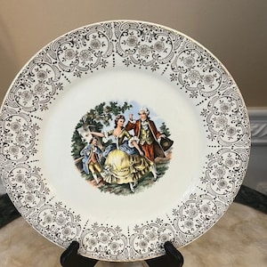 Peut inclure: Une assiette plate en céramique blanche avec un bord doré et un motif floral noir et or. Le centre de l'assiette présente une illustration colorée d'un couple dansant dans un jardin.