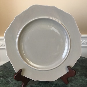 May include: A large, gray ceramic dinner plate with a scalloped edge. The plate is on a wooden stand.
