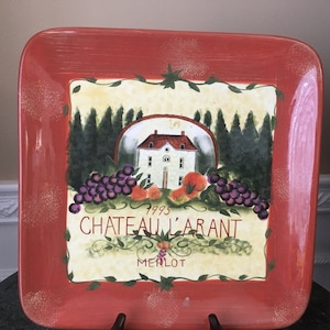 May include: A red ceramic serving platter with a painted design of a house, grapes, and the text "1993 Chateau L'Arant Merlot".