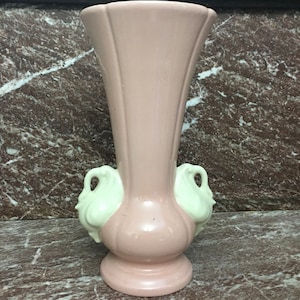 Puede incluir: Un jarrón de cerámica rosa con dos asas de cisne blanco. El jarrón tiene una parte superior acampanada y una base redondeada.