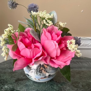 May include: A floral arrangement featuring vibrant pink peonies, small white flowers, and spiky blue accents. The flowers are arranged in a decorative ceramic vase with a landscape design. The arrangement sits on a green marble surface.
