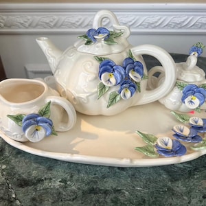 May include: A set of three ceramic mugs with a speckled, matte finish. Each mug features a different color: light blue, cream, and gray. The mugs have a simple, modern design with a rounded shape and a comfortable handle.