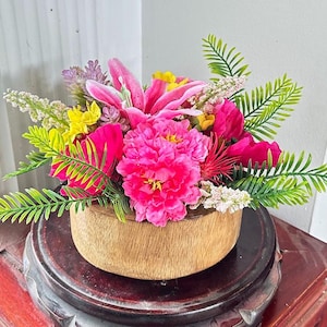 Puede incluir: Un vibrante arreglo floral en un cuenco de madera. El arreglo presenta flores rosas, amarillas y blancas brillantes, con hojas verdes en forma de helecho. El cuenco se asienta sobre un soporte de madera oscura, creando una colorida exhibición.