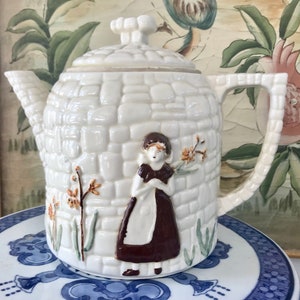 May include: White ceramic teapot with a brick pattern and a girl in a brown dress holding flowers. The teapot has a lid with a knob in the shape of a house.