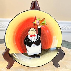 Könnte beinhalten: Ein gelber und roter Keramikteller mit einem Cartoon-Kellner, der ein Tablett mit Essen trägt. Der Kellner trägt einen schwarzen Smoking und eine weiße Fliege. Der Teller hat einen goldenen Rand.