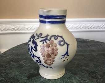 May include: A white ceramic pitcher with a blue and brown grape design. The pitcher has a wide base and a narrow neck.