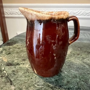 May include: A brown ceramic pitcher with a handle. The pitcher has a glossy finish and a light brown rim. The pitcher is sitting on a green marble table. The pitcher is likely used for serving beverages.