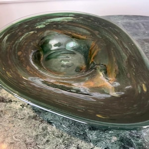 May include: A decorative, teardrop-shaped glass bowl. The bowl is a deep green color with swirling patterns of brown and gold. The glass is transparent, allowing light to pass through. The bowl has a smooth, polished surface.