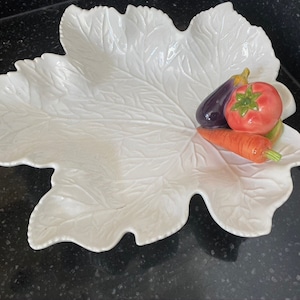 Puede incluir: Plato de servir de cerámica blanca con forma de hoja y textura en relieve. El plato tiene tres pequeñas hendiduras para servir verduras. Una berenjena morada, un tomate rojo y una zanahoria naranja se colocan en las hendiduras.