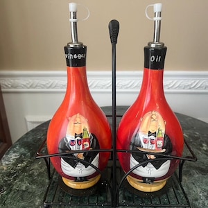 Könnte beinhalten: Ein Set aus roten Keramiköl- und Essigflaschen mit einem schwarzen Metallständer. Die Flaschen haben ein Cartoon-Kellner-Design und sind mit "Essig" und "Öl" beschriftet. Die Flaschen haben eine Höhe von ca. 20 cm.