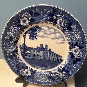May include: A blue and white decorative plate featuring a detailed illustration of a building, possibly a historic mansion, surrounded by floral patterns. The plate has a scalloped edge and the text "WASHINGTON'S MANSION MOUNT VERNON" is visible.