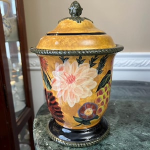 Puede incluir: Un tarro decorativo de cerámica con tapa. El tarro está pintado con un diseño floral en tonos naranja, rosa y verde. La base es negra con un ribete plateado, y la tapa está rematada con un adorno decorativo.