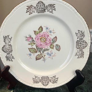 May include: Cream-colored decorative plate with a pink rose and floral design in the center. The plate has a gold rim and gold accents around the edge. The plate is on a black stand.