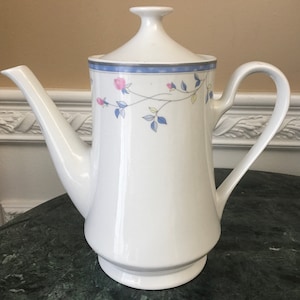 Può includere: Caffettiera in ceramica bianca con un motivo floreale in tonalità blu, rosa e giallo. La caffettiera ha un coperchio e un manico.