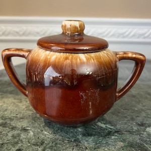 May include: A brown ceramic sugar bowl with a lid and two handles. The bowl has a glossy finish with a gradient effect, transitioning from a lighter brown at the top to a darker brown at the base. The lid has a small knob.
