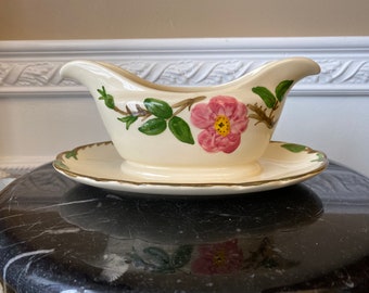 May include: A white gravy boat with a pink rose and green leaves design. The boat has a gold trim and comes with a matching saucer.