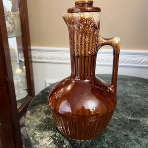 Puede incluir: Una jarra de cerámica marrón con tapa. La jarra tiene una base bulbosa, un cuello alto y acanalado, y un asa. La parte superior y la tapa tienen un esmalte más claro y moteado. La jarra es probablemente vintage y adecuada para servir bebidas.