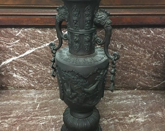 May include: A black, ornate, metal vase with intricate carvings and two handles. The vase has a round base and a tall, slender body.