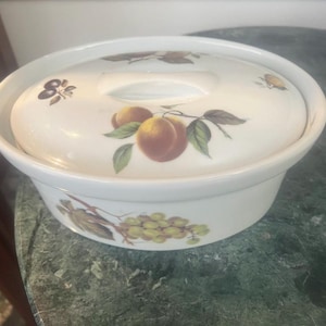 Op de afbeelding: Een witte keramische ovenschaal met bijpassend deksel, voorzien van een decoratief patroon van perziken, groene druiven en donkerpaarse pruimen met groene bladeren, geschilderd op de buitenkant van de kom en de bovenkant van het deksel.