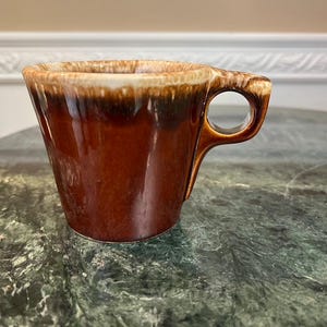 Puede incluir: Una taza de cerámica marrón con un diseño de asa único. La taza tiene una abertura ancha y un exterior marrón oscuro que se desvanece a un marrón más claro y crema en el borde. Está sobre una superficie de mármol verde.