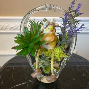 以下が含まれることがあります： 取っ手付きのバスケット型の透明なガラスの花瓶。緑の多肉植物、紫の花、小さな黄色の鳥の置物が入っています。