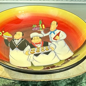 Könnte beinhalten: Eine große, runde Servierschüssel aus Keramik mit einem farbenfrohen Design, das drei Cartoon-Köche zeigt. Die Köche halten verschiedene Lebensmittel in der Hand, darunter einen Teller Pasta, ein Tablett mit Gebäck und eine Flasche Wein. Die Schüssel hat einen gelben, orangen und roten Farbverlauf im Hintergrund.