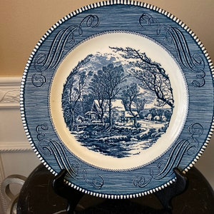 Puede incluir: Plato de cena de cerámica azul y blanco con una ilustración azul y blanco de una escena de invierno con una granja y árboles. El plato tiene un borde azul y blanco con un patrón decorativo.