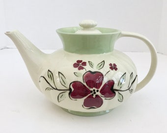 May include: A white teapot with a green lid and floral design. The teapot features a large, red flower with black accents and smaller red flowers with green leaves.
