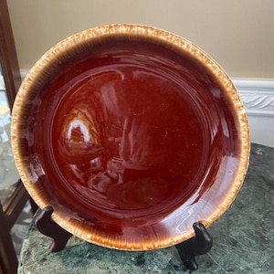 Puede incluir: Un plato redondo de cerámica esmaltada con un centro marrón rojizo profundo y un borde texturizado de color beige. El plato se exhibe sobre un soporte negro, mostrando su acabado brillante y estética vintage. Una pieza clásica.