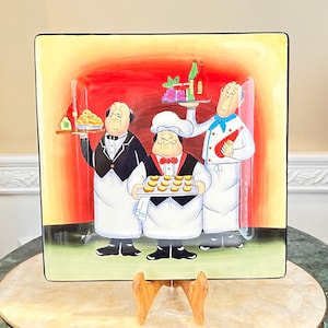 Könnte beinhalten: Eine Keramik-Servierplatte mit einer farbenfrohen Illustration von drei Köchen. Die Köche tragen weiße Schürzen und halten Tabletts mit Essen. Der Hintergrund ist ein roter und gelber Farbverlauf.