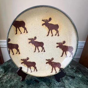 Puede incluir: Un plato redondo de cerámica con fondo crema y siluetas de alces marrones. El plato tiene un borde verde oscuro y se exhibe sobre un soporte negro. Este plato decorativo es perfecto para servir comida o como pieza decorativa.