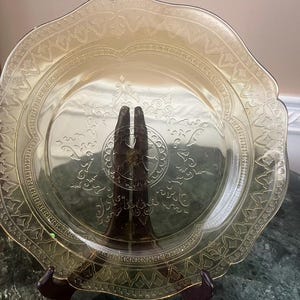 May include: An antique amber glass plate featuring a scalloped rim and detailed embossed floral and geometric designs. The plate is presented on a dark brown stand, highlighting its delicate craftsmanship. A decorative item for serving or display.