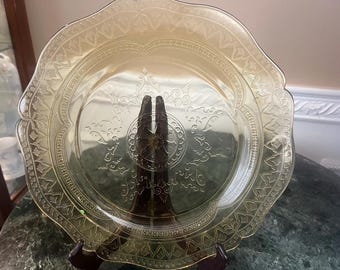 May include: A vintage amber-colored glass plate with a scalloped edge and intricate embossed floral and geometric patterns. The plate is displayed on a dark brown stand, showcasing its delicate design. A decorative piece for serving or display.