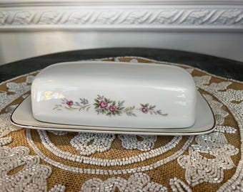 Könnte beinhalten: Weißes Porzellan-Buttergericht mit rechteckiger Basis und gewölbtem Deckel. Das Gericht ist mit einem floralen Muster mit rosa Rosen und grünen Blättern verziert. Die Ränder sind mit einem goldenen Band versehen. Ein klassisches Stück zum Servieren von Butter.