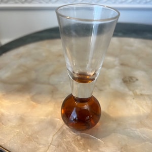 Può includere: Un bicchierino da shot in vetro trasparente con una base rotonda di colore ambrato. Il bicchiere è vuoto e si trova su una superficie bianca.