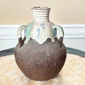 Puede incluir: Un jarrón de cerámica marrón con dos asas y un esmalte blanco. El jarrón tiene una superficie rugosa y texturizada y un aspecto rústico único.