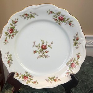 May include: White porcelain plate with a scalloped edge and gold trim. The plate is decorated with a floral pattern of pink roses and green leaves. The plate is displayed on a black stand.
